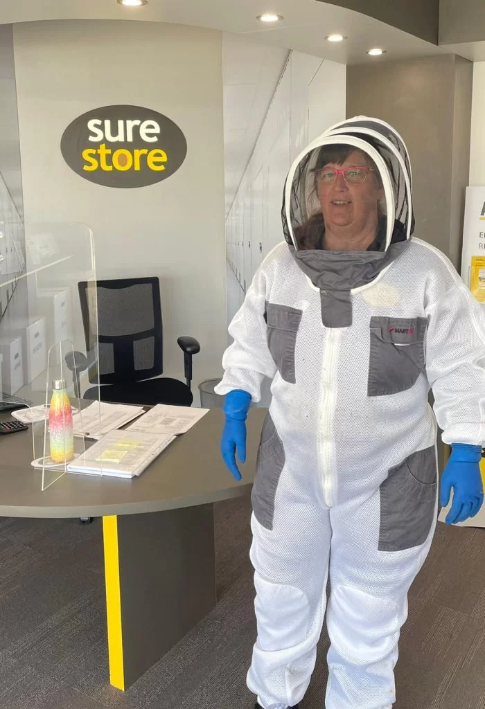 CLAIRE-BEEKEEPER-700×1024 A woman wearing a protective beekeeping suit and veil stands at a reception desk in an office with a logo that reads "sure store." The setup includes a chair and paperwork on the desk.