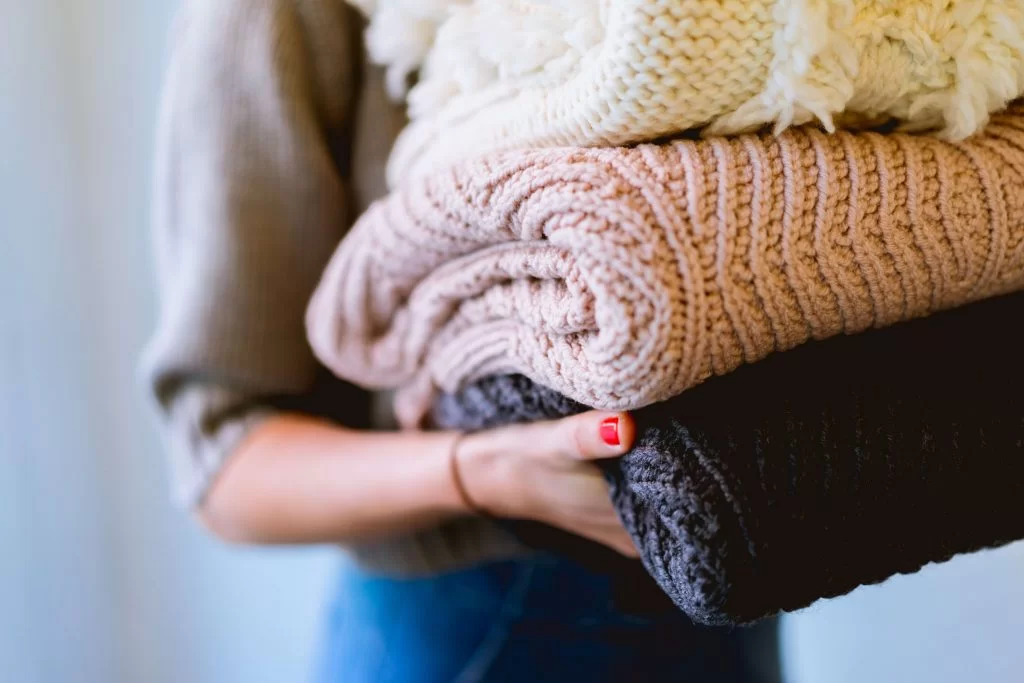 folded-clothes-1024×683 A person holding three neatly folded blankets in neutral tones. The blankets are textured, with one in light cream, one in soft pink, and one in dark gray. The person's arm is visible, showing a hand with red nail polish.