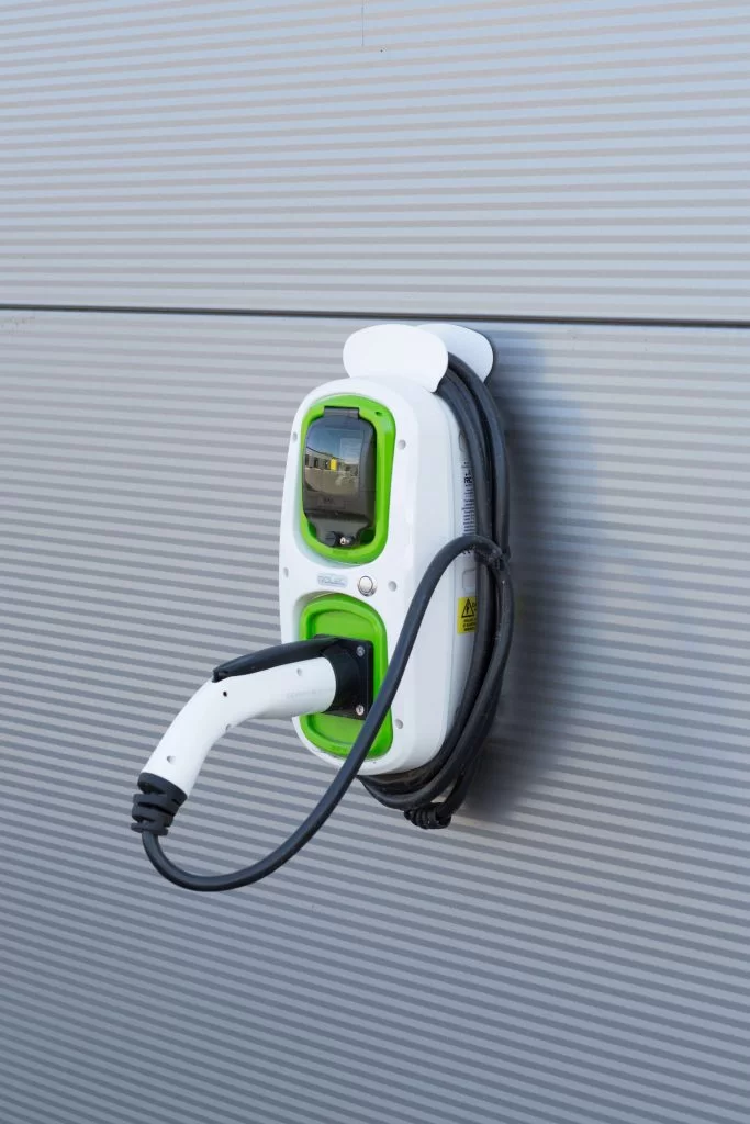 surestore-bolton-230518-001_-683×1024 An electric vehicle charging station mounted on a wall, featuring a digital display and a cable with a connector hanging down. The station has a white finish with green accents.