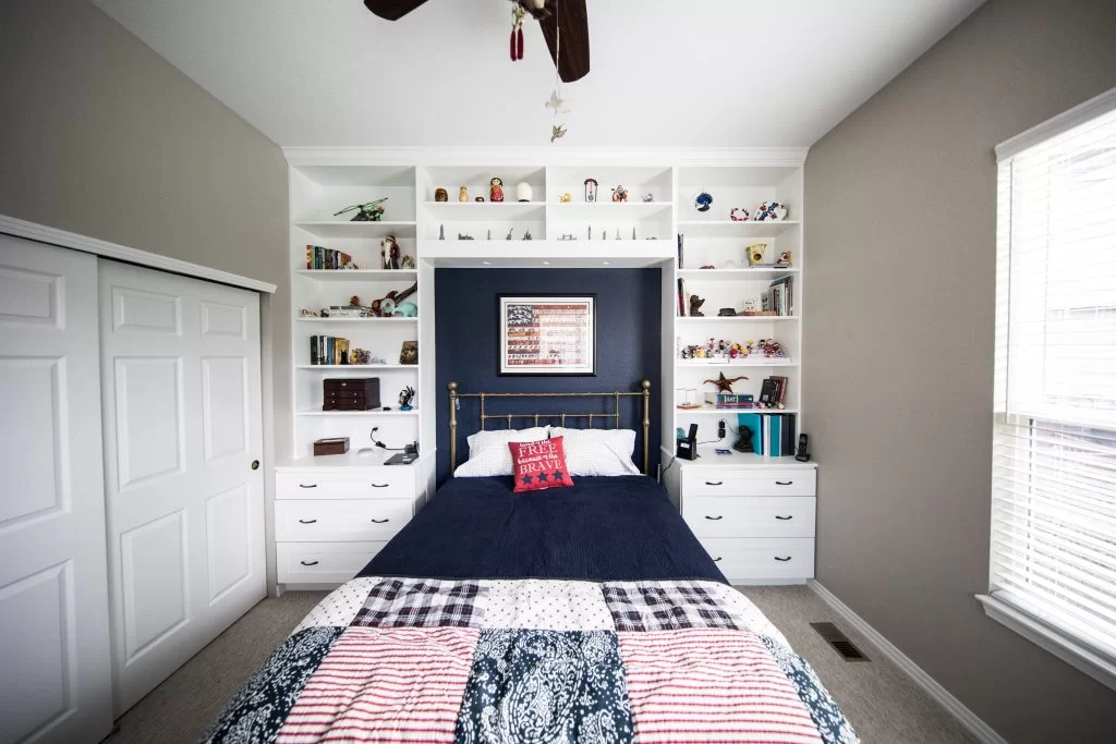 use-shelving-1024×683 A neatly arranged bedroom featuring a dark blue bedspread on a bed with white pillows. Behind the bed, there is a blue wall with shelves displaying various items, and a framed American flag. White closet doors are visible on the left.