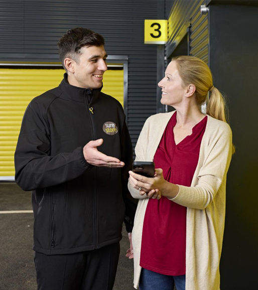 how-to-book-surestore A man in a black jacket and a woman in a red top and cream cardigan are engaged in conversation. They stand beside a storage unit with yellow doors, with a number 3 visible on the wall.