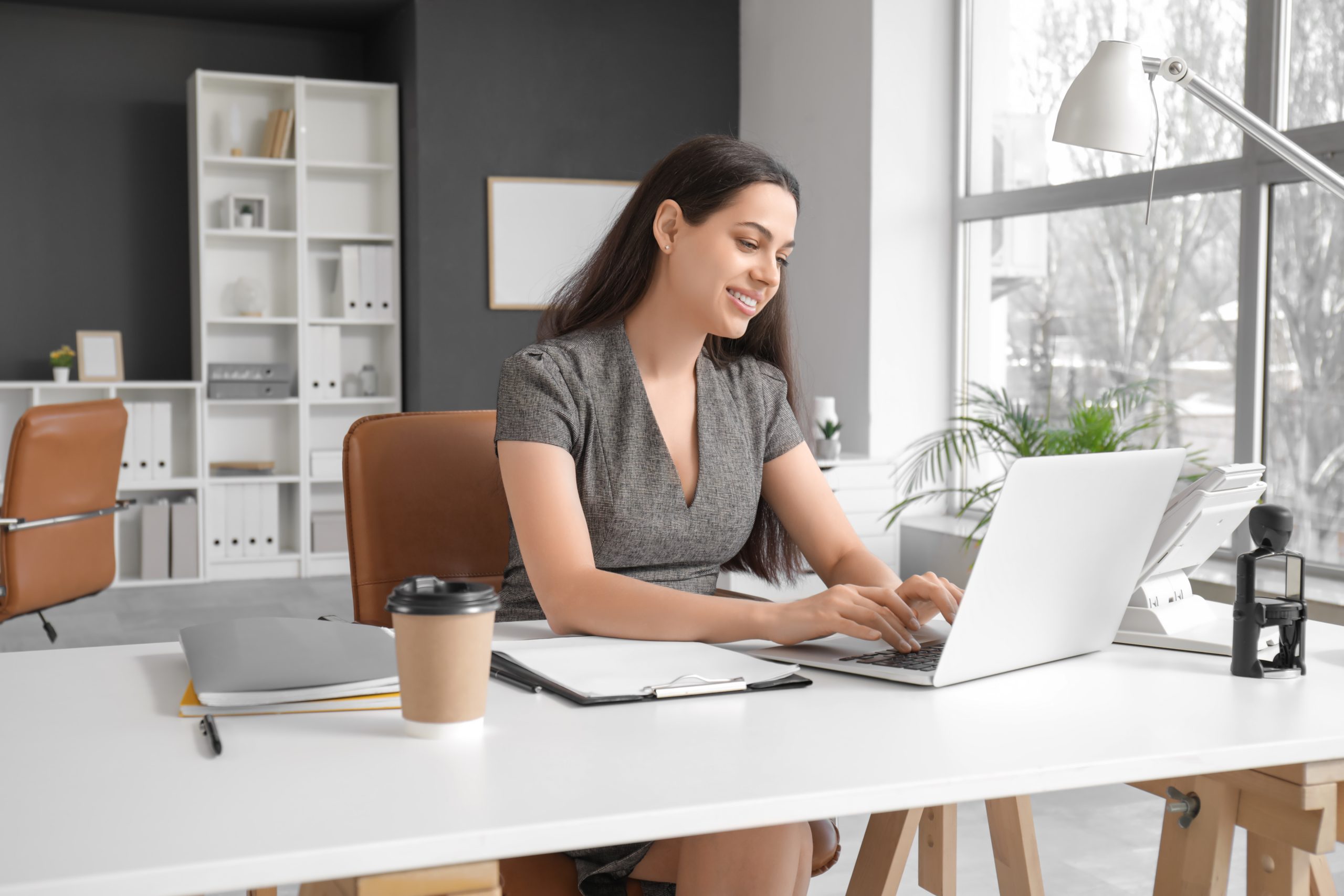 A woman sitting at a desk in a modern office, using a laptop. She is smiling while typing, with a notebook and a coffee cup on the table. The office has large windows and light-colored furniture.