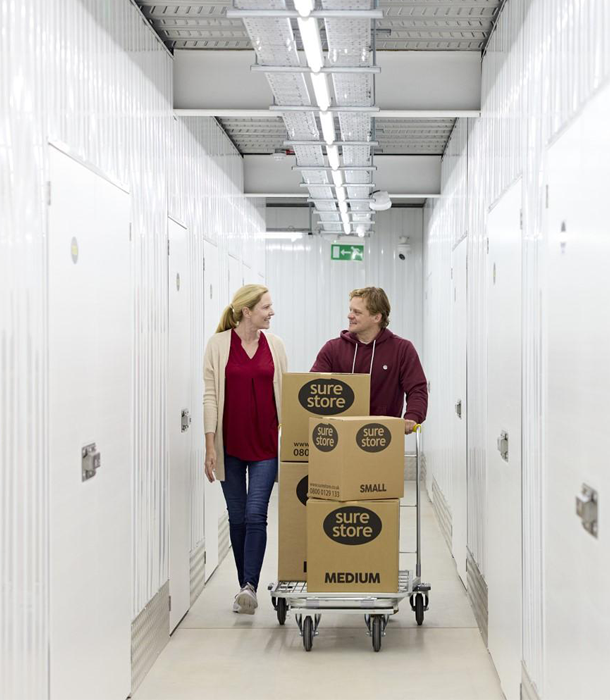 why-store-with-us A woman and a man walking together in a storage facility hallway, with the man pushing a cart stacked with cardboard boxes labeled "sure store" in various sizes.